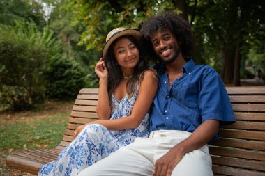 A young interracial couple, he is African,she is Asian, they are sitting on a bench in a public park and smiling