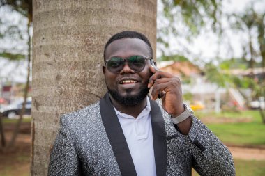 Young smiling black man on vacation, leaning against a tree makes a call.