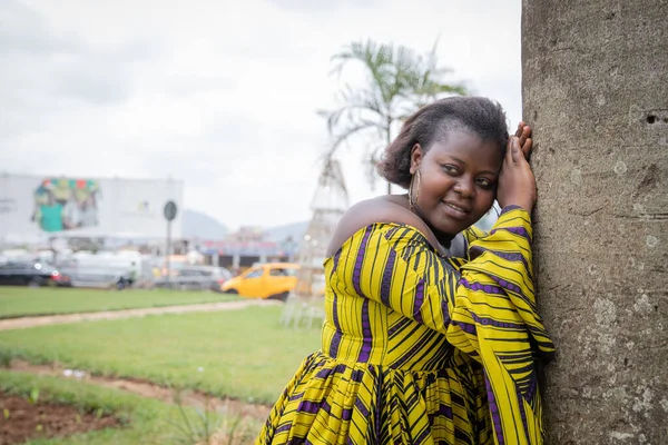 Young serious African woman leaning against a tree gets her picture taken.