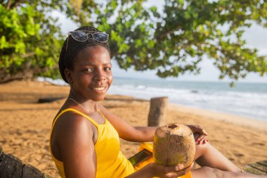Beautiful girl enjoys her beach vacation sitting on the sand drinking coconut milk.