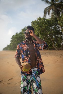 Young man by the sea with a coco nut in hand smiles as he looks at the horizon.