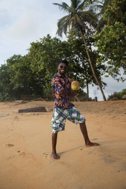 Barefoot on the sand, young man in sunglasses smiles and holds a coconut ready to be tasted.
