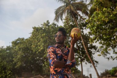 Frame of a young man looking into the distance and holding a large coconut with a straw.