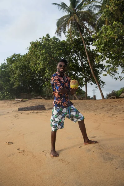 Barefoot on the sand, young man in sunglasses smiles and holds a coconut ready to be tasted.