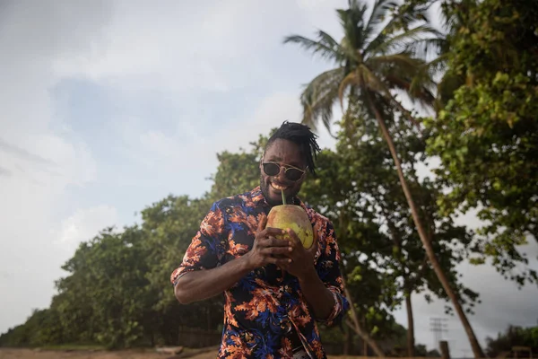 A gentleman smiles while enjoying coco milk from the coconut he is holding.