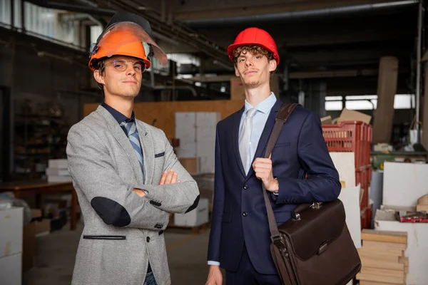 Portrait of two engineers in the factory, successful people elegantly dressed wearing protective helmets