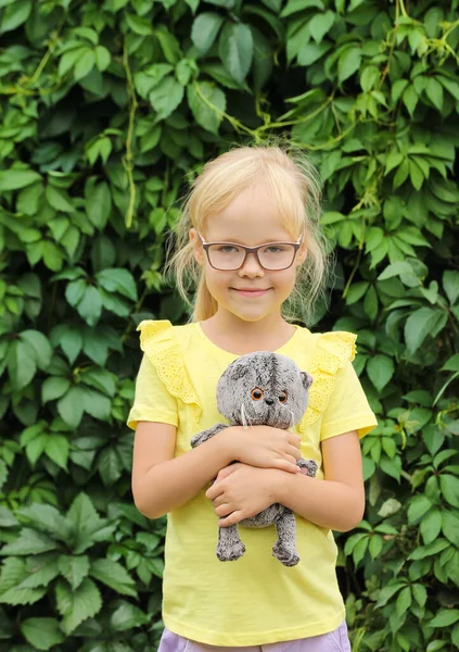 Fille blonde en lunettes et un T-shirt jaune avec un chaton jouet préféré bébé Basique dans le jardin sur fond de verdure — Photo