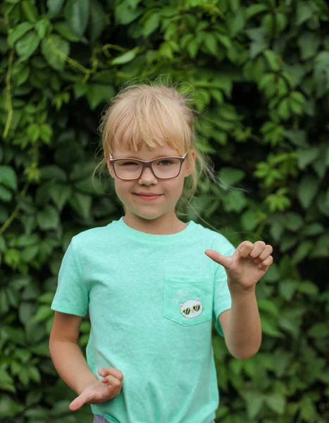 Un enfant mécontent avec des lunettes dans un t-shirt à la menthe dans le jardin sur fond de feuilles vertes — Photo