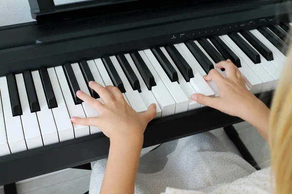 Маленькая блондинка играет на цифровом пианино в гостиной, крупным планом сзади. Ребенок учится играть на музыкальном инструменте. Раннее музыкальное образование для детей. — стоковое фото