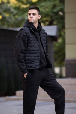 Siyah kostümlü ve yelekli bir adam şehirde poz veriyor.