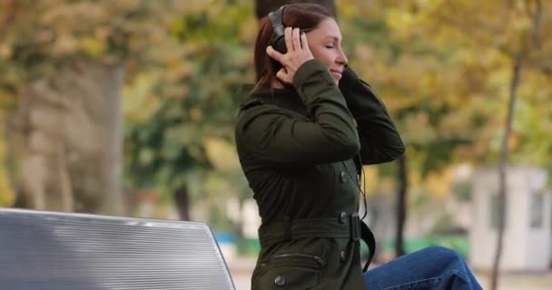 Kvinnan lyssnar ljudböcker bär hörlurar sitter på bänken i en park — Stockvideo