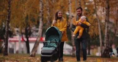 Genç bir aile sonbahar parkında bebek arabasıyla yürüyor.