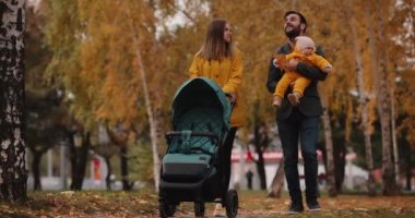Genç bir aile sonbahar parkında bebek arabasıyla yürüyor.