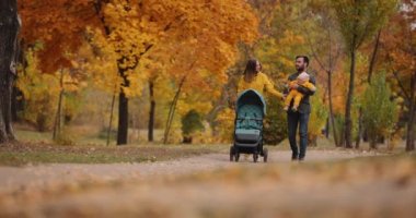 Genç bir aile sonbahar parkında bebek arabasıyla yürüyor.