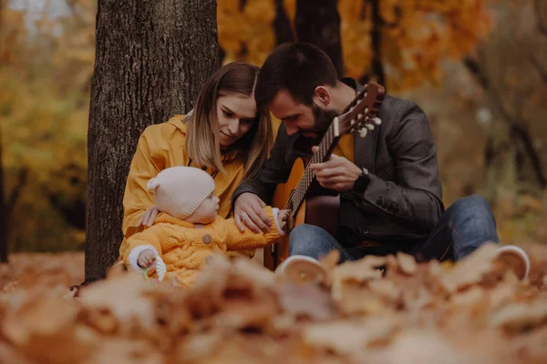 Onnellinen nuori perhe, jolla on pieni vauva syksyllä puistossa, isä soittaa kitaraa — kuvapankkivalokuva