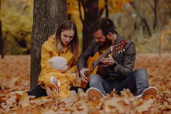 Glad ung familj med liten bebis i höstparken, pappa spelar på gitarr — Stockfoto