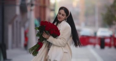 Mutlu kadın şehirde çiçekleri taşıyor ve kokluyor.