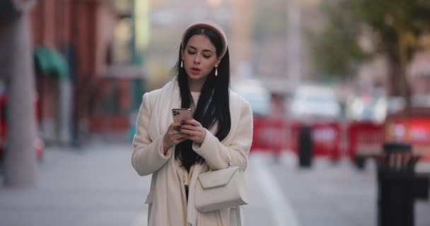 Молода стильна жінка дивиться в смартфон, що йде містом — стокове відео