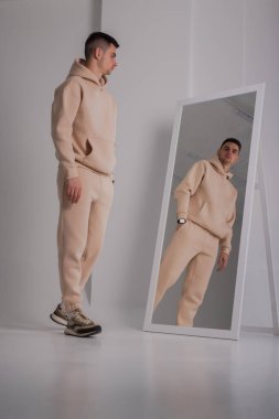 Stüdyoda bej kapüşonlu kostüm giyen bir adam aynanın yanında poz veriyor.