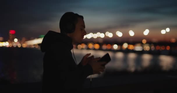 Człowiek wybiera muzykę ze swojej listy odtwarzania stojąc na nabrzeżu miasta — Wideo stockowe