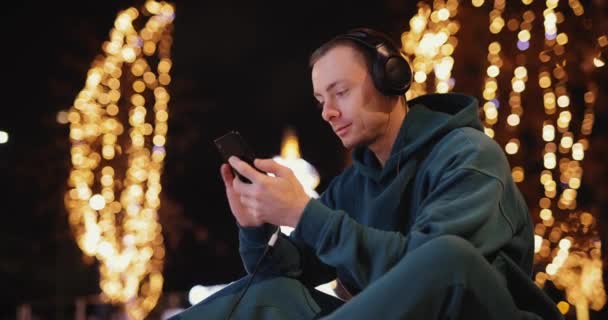 Νεαρός που χρησιμοποιεί το τηλέφωνο σε ακουστικά στην πόλη το βράδυ — Αρχείο Βίντεο