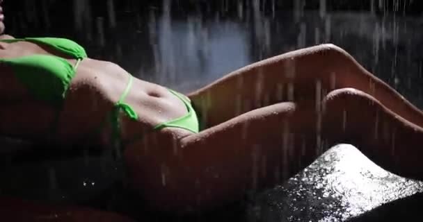 Сексуальная женщина в бикини лежит на полу под дождем в черной студии — стоковое видео