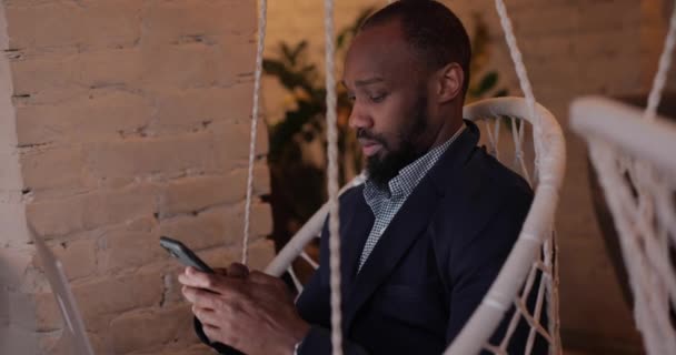 Uomo d'affari africano che utilizza smartphone seduto in amaca nel caffè — Video Stock