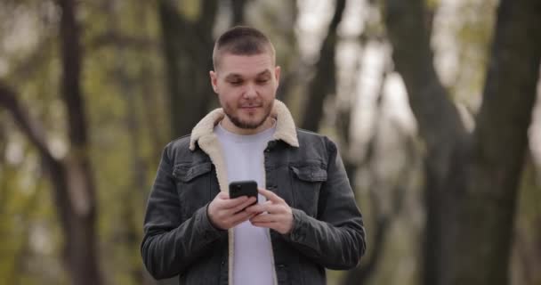 El hombre sonriente de cámara lenta usa un teléfono inteligente caminando por el parque — Vídeo de stock