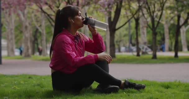 Sporty woman drinking water from shaker sitting in the park — Stock video