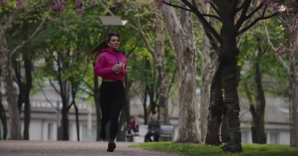 Pretty sporty woman jogging in the summer park with earbuds listening to music — Wideo stockowe