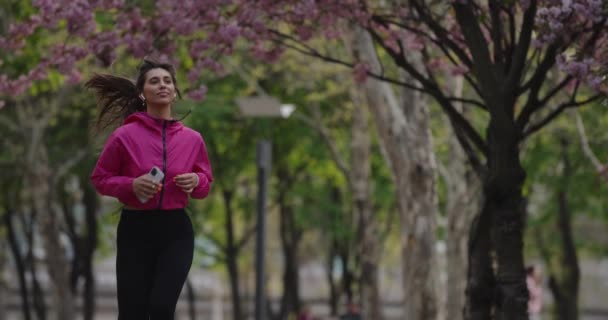 Pretty sporty woman jogging in the summer park with earbuds listening to music — Stockvideo