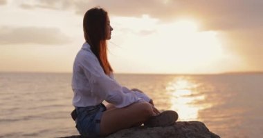 Genç bir kadın gün batımını seyrediyor. Tepede oturuyor, ağır çekimde.