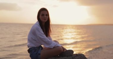 Gün batımında deniz kenarında oturan genç bir kadın, yavaş çekimde taş gibi hareketler yapsın.
