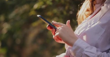 Hands of young unrecognizable woman using phone sitting in the park closeup
