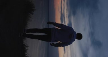 Silhouette of man walking on a hill at the evening looking at beautiful sea view, vertical video