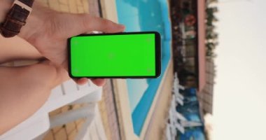 POV male hand holding smartphone with empty green screen chroma key relaxing near pool, vertical video