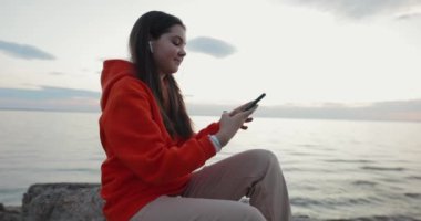 Gün batımını akıllı telefondan fotoğraflayan genç bir kız deniz kenarında taşın üzerinde oturuyor.