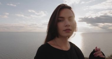 Muhteşem deniz ve gökyüzü manzarasının üzerindeki tepede duran kameraya bakan kadının yavaş hareket eden portresi.