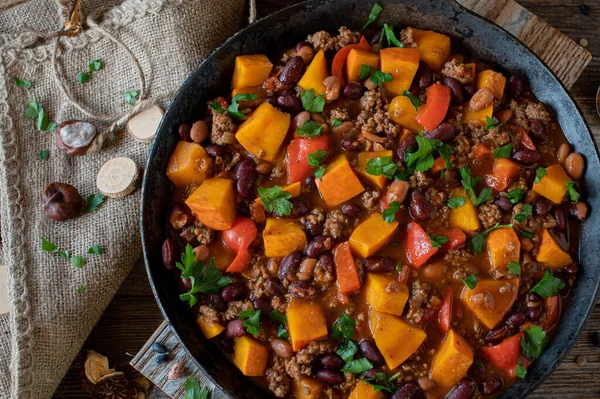 Homemade stew with pumpkin and ground beef. Homemade stew with ground beef, pumpkin and kidney beans cooked with vegetables such as tomato, bell peppers, garlic and herbs. Served in rustic cast iron skillet on wooden table. Overhead view.