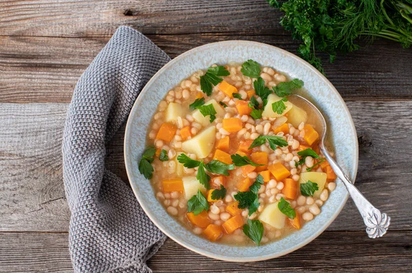 Vegan or vegetarian bean soup with root vegetables. Fresh and homemade cooked white beans soup, vegan cuisine, with potatoes, carrots and root vegetables. Served in a  bowl with spoon on rustic and wooden table. Overhead view