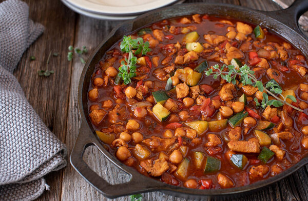 Chickpea stew with chicken breast and mediterranean vegetables such as tomatoes, zucchini, onions, red bell peppers and garlic. Served in a delicious tomato sauce in a rustic cast iron pan on wooden background from above. 