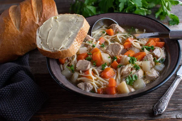 Bowl with soup and bread. Fresh and homemade cooked chicken noodle soup served with baguette and butter in a rustic enamel bowl on wooden table.