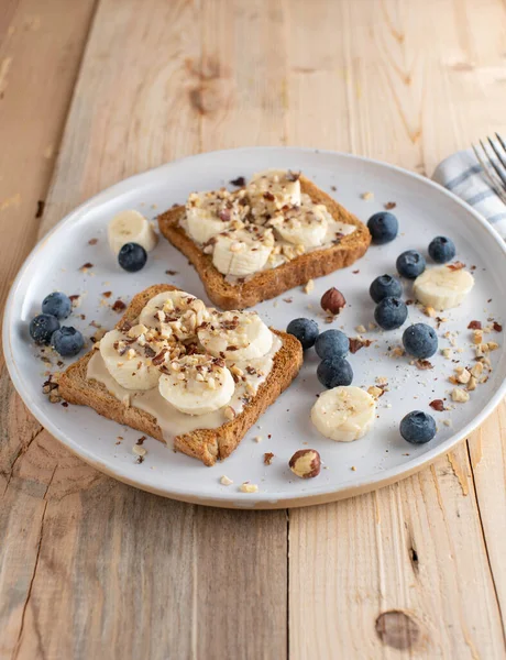 Clean eating breakfast plate with whole grain toast topped with almond butter, banans and hazelnuts. Served with fresh berries on a plate isolated on wooden table with copy space