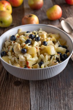 Elmalı, muzlu, sarı kivili ve yabanmersinli taze ve ev yapımı meyve salatası. Koyu ahşap masa arkasında beyaz bir kasede servis edilmiş. Kopyalama alanı olan dikey resim