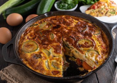 Sebzeli Frittata. Fırın, kabak, havuç, dolma biber, domates ve soğanla pişirilmiş. Ahşap masa arka planında ütü tavasında servis edilmiş.