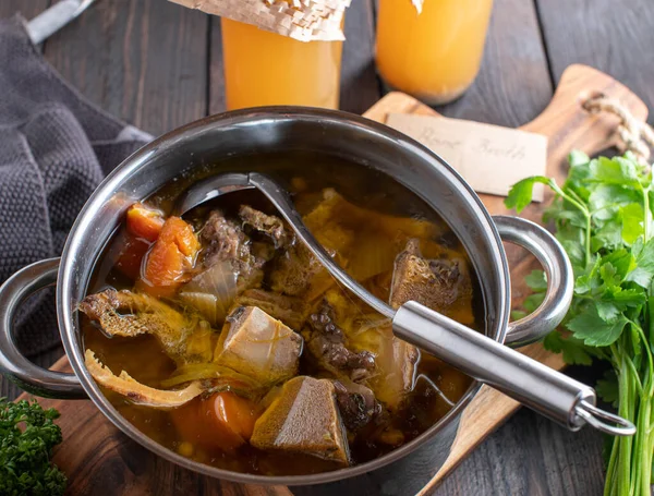 国产肉汤 用有机牛肉骨头 根茎蔬菜和香草烹调而成 放在一个锅里 锅里有勺子和罐子 放在粗糙的木桌背景上 — 图库照片