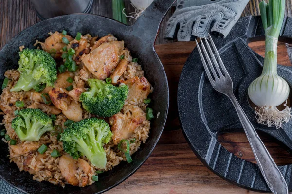 Healthy fitness meal with pan fried and chopped chicken breast, brown rice and broccoli. Served in a rustic iron pan on wooden table background with dumbbell. Top view