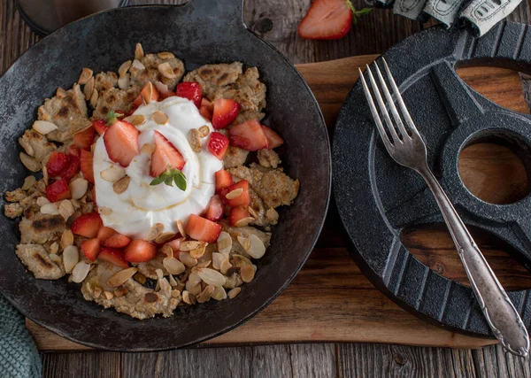 Healthy and delicious homemade fitness meal with a fresh shredded oatmeal pancake, fresh strawberries, protein quark, roasted almonds and maple syrup. Served in a rustic iron pan on wooden table background with dumbbell