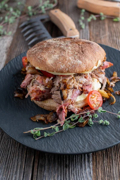 Burger with pulled pork, cheese, tomatoes and roasted onions on a plate on rustic and wooden table background. Traditional american barbecue sandwich.