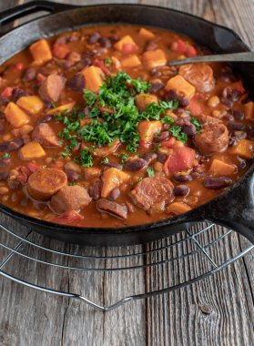 Baharatlı yahni, Meksika ya da Tex Meksika usulü sucuklu, tatlı patatesli, fasulyeli ve sebzeli. Dökme demir tavada servis edilir. Ahşap masa arkasında izole edilmiş..